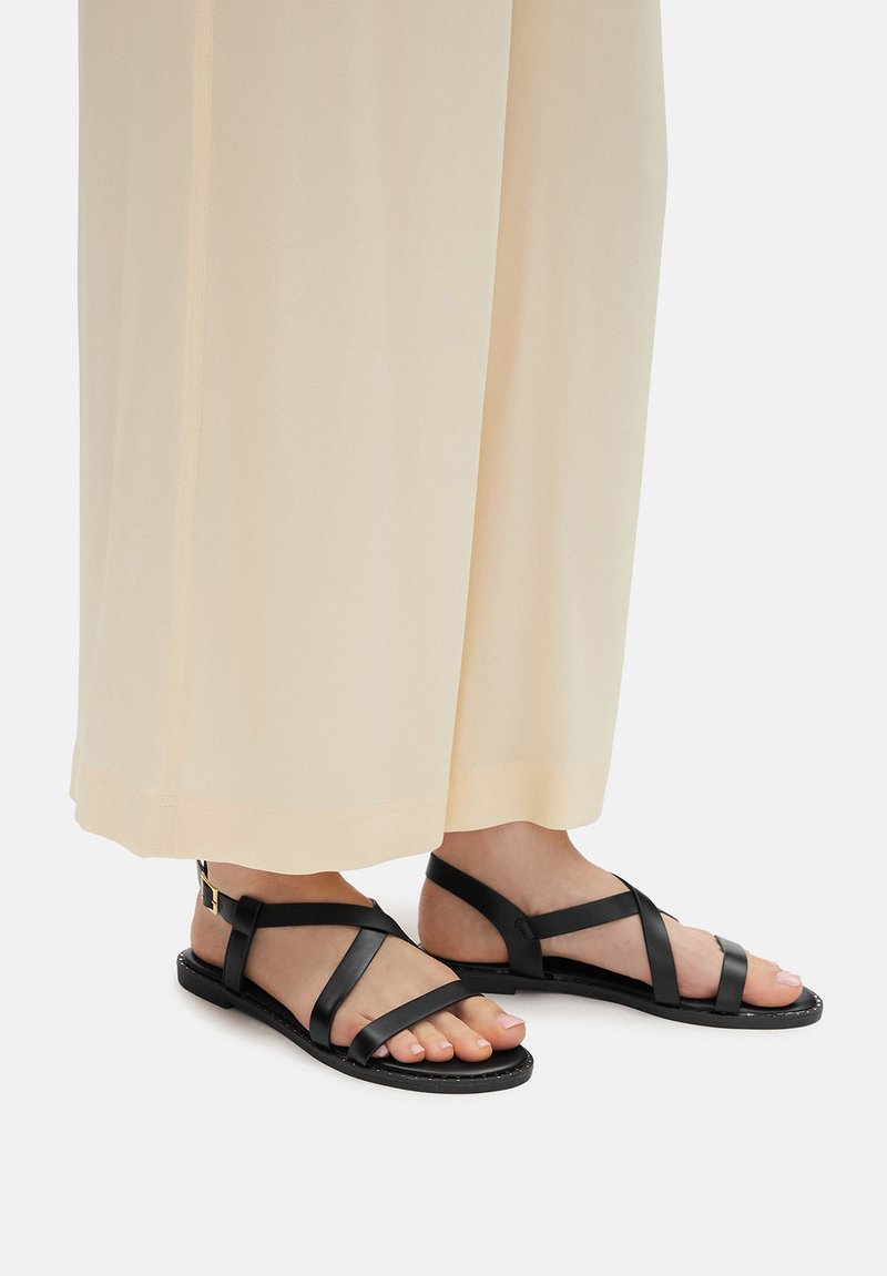 Czarne skórzane sandały z krzyżującymi się paskami i płaską podeszwą, noszone z jasnymi, szerokimi spodniami, podkreślające minimalistyczny design.