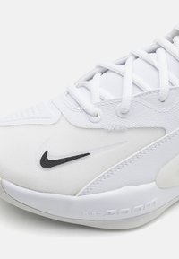 Hvide atletiske sko med en tekstureret overdel, glatte læderdetaljer og sort Nike-logo. Har snørebånd og polstret sål med "Nike Zoom" detalje.
