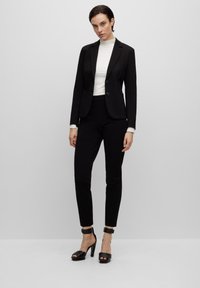 Zwarte op maat gemaakte blazer met twee knopen, gecombineerd met een strakke zwarte broek en een witte top met hoge hals. Zwarte sandalen met hoge hakken maken de look compleet.