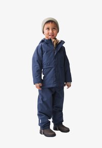 Navy waterdichte jas met een capuchon, bruine gebreide beanie, beige binnenlaag en bijpassende broek. Boots met elastische manchetten bij de enkels.