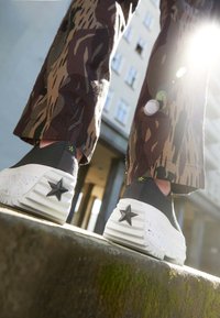 Svarta slip-on skor med en texturerad ovandel och vit räfflad sula, med en svart stjärndetalj på hälen. Mönstrade bruna byxor syns.