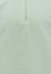 Tissu vert clair avec une fermeture éclair argentée verticale partiellement fermée au centre, montrant des coutures détaillées autour de la base de la fermeture éclair.