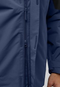 Veste imperméable bleu marine avec une fermeture éclair robuste, un tissu texturé et des accents en velcro sur les côtés pour un ajustement réglable.