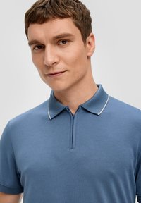 Niebieska koszulka polo z zamkiem przy szyi, krótkimi rękawami i kontrastowym białym wykończeniem kołnierzyka. Materiał wydaje się gładki z lekką teksturą.