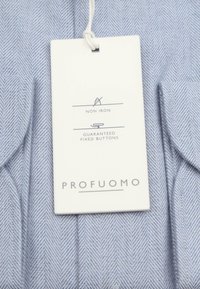 Lichtblauwe visgraatstof met een wit label waarop staat dat het non-iron is, met gegarandeerde vaste knopen en het merk Profuomo.