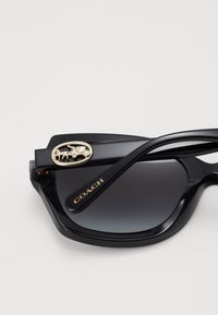 Lunettes de soleil en plastique noir avec des verres dégradés, présentant un emblème doré d'un cheval et d'une voiture sur la branche, ainsi que la marque "COACH".