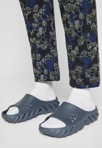 Grå slip-on sandaler med en strukturerad sula. Har öppen tå-design och ventilationshål, bärs med vita strumpor och mönstrade byxor.