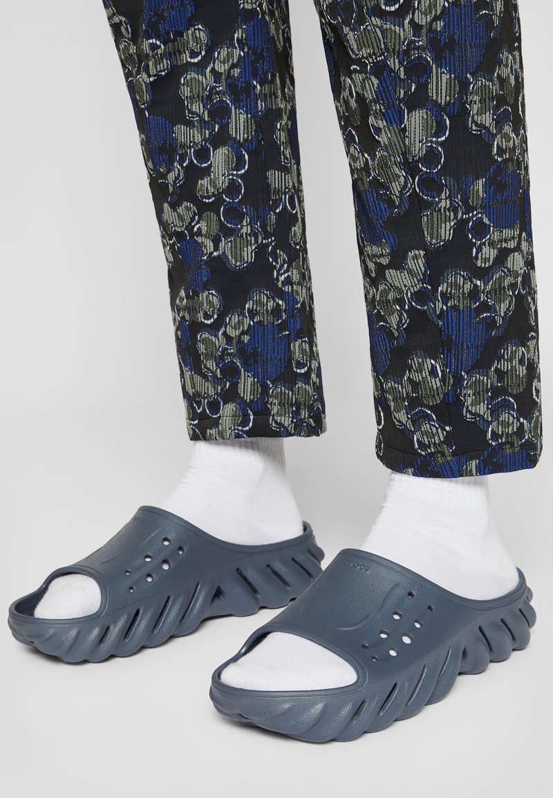 Grå slip-on sandaler med en strukturerad sula. Har öppen tå-design och ventilationshål, bärs med vita strumpor och mönstrade byxor.