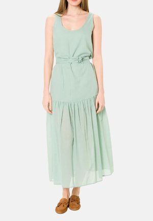 Donna che indossa un vestito maxi verde menta senza maniche con cintura e gonna arricciata, e pantofole mocassino marroni, in piedi su sfondo bianco.