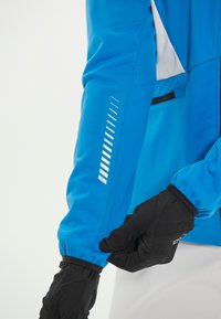 Blaue Softshelljacke mit elastischen Bündchen; besitzt ein weißes Streifenmuster und eine Reißverschlusstasche. Schwarze Handschuhe passen den Ärmel an.