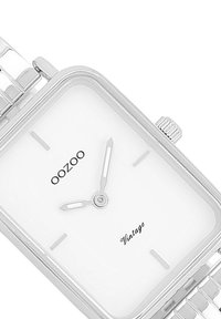 Montre rectangulaire en argent avec un cadran blanc, design minimaliste, aiguilles métalliques fines et nom de la marque 'OOZOO' avec détail de texte 'vintage'.
