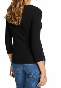 Frau mit langen blonden Haaren, die ein schwarzes langärmliges, geripptes Oberteil und blue Jeans trägt, von hinten gezeigt, mit Armbändern am rechten Handgelenk.