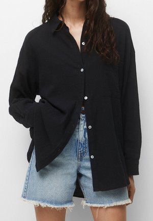 Woman wearing loose black button-up shirt with one hand in pocket and light blue frayed denim shorts against white background.