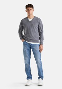 Szary sweter w dekolcie w serek o dzianinowej strukturze, włożony na białą koszulę. Połączony z jasnoniebieskimi dżinsami i białymi trampkami. Gładkie tło.