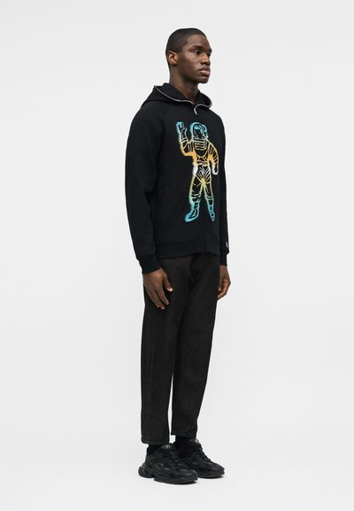 Άνδρας που στέκεται, φορώντας μαύρο φούτερ με φερμουάρ και πολύχρωμο σχέδιο αστροναύτη, μαύρο παντελόνι και μαύρα αθλητικά παπούτσια, μπροστά σε απλό φόντο.