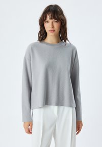 Maglione grigio a maniche lunghe con collo rotondo, texture a costine, vestibilità rilassata e orlo corto, abbinato a pantaloni bianchi.
