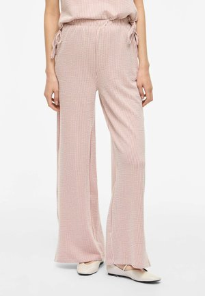 Kobieta nosząca różowe i białe paskowane spodnie z wysokim stanem i szerokimi nogawkami, z wiązaniami po bokach, w zestawieniu z beżowymi balerinami.