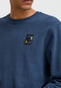 Marineblå genser med rund hals, med en brodert "BORN LUCK" design og en stilisert figur som holder en sko.