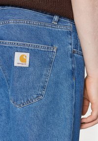 Blue denim jeans featuring a back pocket with a square logo patch in yellow and white, stitched detailing, and a classic cut.