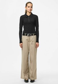 Melnas, garām piedurknēm polo krekls ar ribotu tekstūru un trīs pogu aizdari, kopā ar bēšām platām biksēm un melnu jostu.