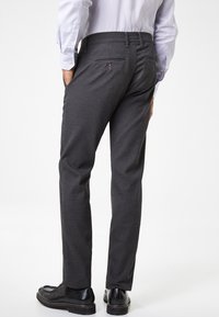 I pantaloni grigi intrecciati presentano un taglio dritto, due tasche posteriori con bottoni e sono abbinati a scarpe nere. Il tessuto ha una texture sottile.