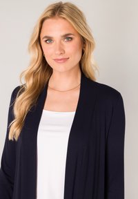 Marine gebreide cardigan met een open voorkant, gedragen over een witte top. De stof lijkt glad met een lichtgewicht textuur.