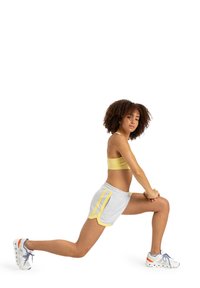 Pantalones cortos deportivos grises con rayas amarillas a los lados, combinados con un sujetador deportivo amarillo. El sujeto está realizando un ejercicio de zancada. Zapatillas blancas con acentos.