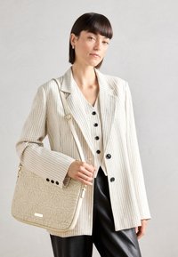 Sac en bandoulière beige matelassé avec motif de roses, associé à un blazer crème rayé et un pantalon noir, avec des boutons noirs et une coupe décontractée.