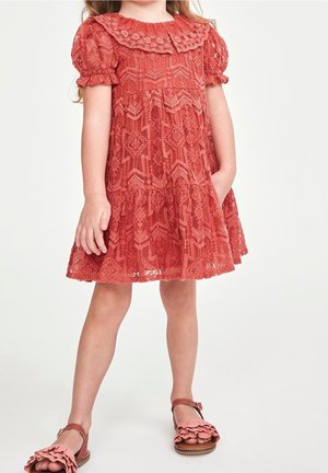 Robe en dentelle corail avec de courtes manches bouffantes, col festonné, jupe à volants et un motif floral texturé. Sandales brunes avec des accents froncés.