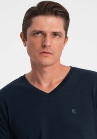 Námořnická modrá tričko s výstřihem do V vyrobené z měkké tkaniny, s malým logem na hrudi a hladkou, nezdobenou texturou.