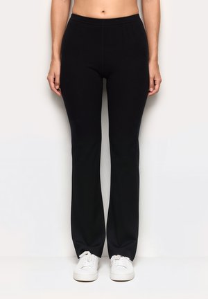 Schwarze, dehnbare Hosen mit einem taillierten Bund und weit geschnittenen Beinen, aus weichem Material. Kombiniert mit weißen Turnschuhen, was einen lässigen Look erzeugt.