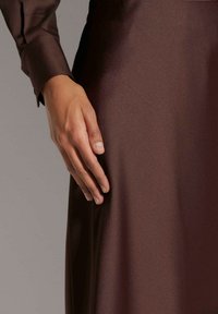 Bruine satijnen stof met een gladde textuur, die elegant valt. Een hand raakt de stof licht aan, waardoor de zachtheid en glans worden benadrukt.