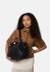 Giovane donna con capelli ricci che indossa una camicia marrone e una gonna chiara, mentre tiene in mano una grande borsa in pelle intrecciata nera con fibbie dorate.