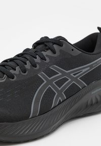 Svart sportskor med en texturerad mesh-överdels, platta svarta snören och en flexibel sula; har en grå logo och ett subtilt mönster på sidan.