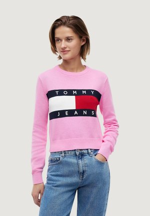Frau trägt einen rosa Tommy Jeans Pullover mit Logo und hellblaue Jeans, steht mit einer Hand in der Tasche vor einfarbigem Hintergrund.