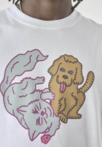 Karikaturstil blauer Kater, der mit einem braunen Hund auf einem weißen T-Shirt spielt. Der Kater liegt auf dem Rücken und hat eine rosa Pfote sichtbar; der Hund streckt die Zunge heraus.