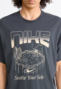 Sort bomulds T-shirt med et gyldent grafisk tryk, der har stor "NIKE" tekst, en illustreret gennemblødt sko og teksten "Soothe Your Sole".