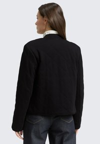 Czarna pikowana kurtka z wzorem w romby, z niskim tyłem i klasycznym kołnierzem. Noszona na ciemnych dżinsach.