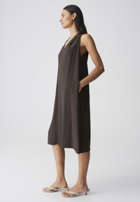 Woman standing in profile wearing a sleeveless dark brown midi dress and beige low-heeled sandals with hands in dress pockets.