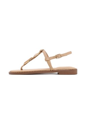 Sandalia plana color beige con suela fina, diseño de tira en T, pequeña hebilla dorada en la correa del tobillo y estilo minimalista sobre fondo blanco.