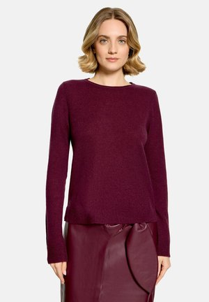 Frau mit kurzen blonden Haaren, die einen langärmeligen lila Pullover und einen passenden lila Lederrock mit Drapierung trägt.