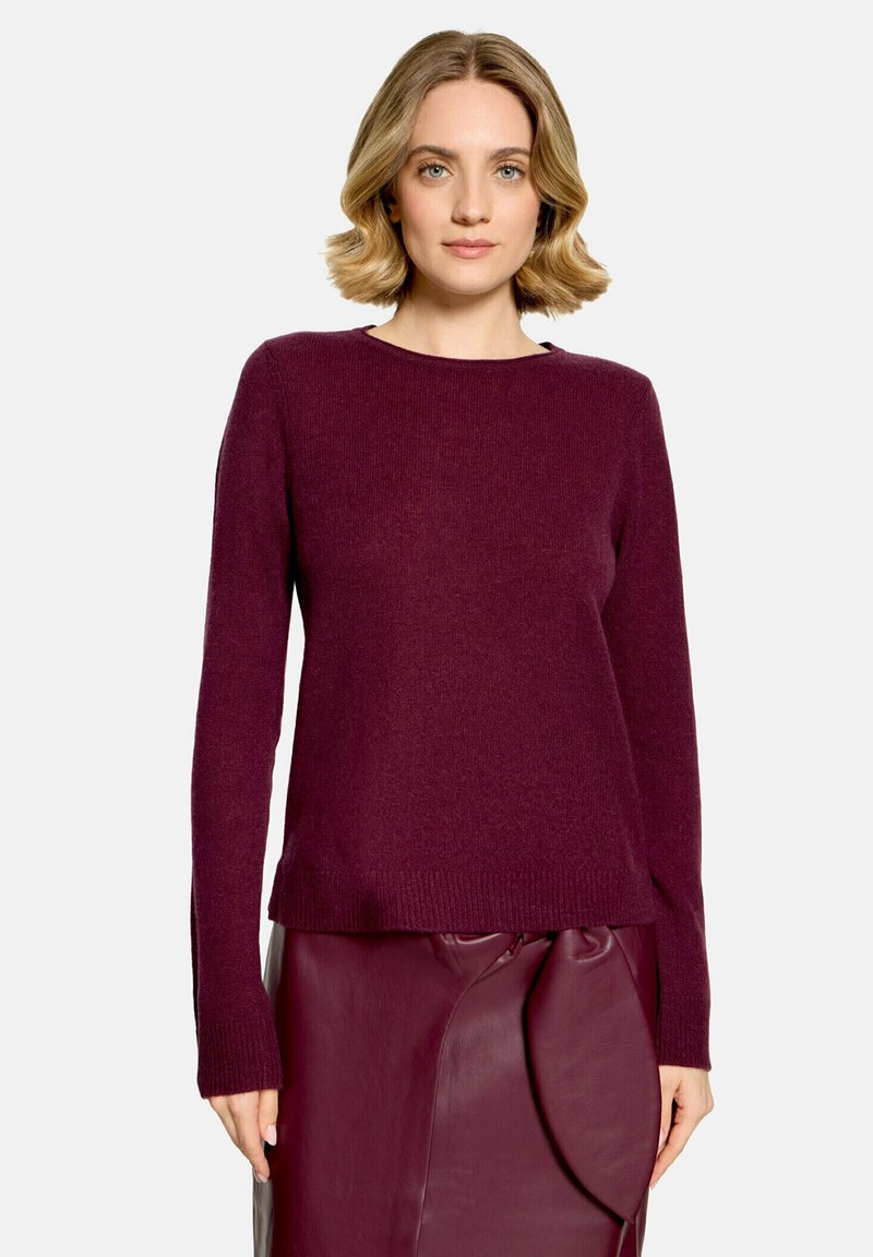 Femme aux cheveux blonds courts, portant un pull violet à manches longues et une jupe en cuir violette assortie avec un détail drapé.