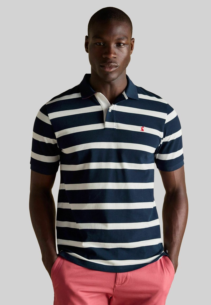 Jūras zils un balts svītrains polo krekls ar īsām piedurknēm, ar apkakli un mazu sarkanu logotipu uz krūts, kombinēts ar rozā biksēm.