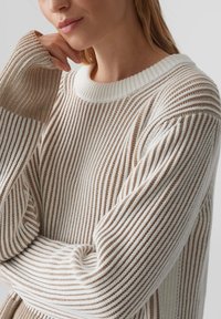 Ein gestrickter Pullover mit wechselnden beige- und weißen Streifen, einer gerippten Textur und einem weiten Rundhalsauschnitt, abgebildet am Oberkörper eines Modells.