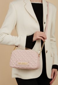 Borsa a tracolla rosa trapuntata con hardware dorato, logo impresso e una tracolla a catena. Presenta una forma rettangolare e una texture in pelle morbida.