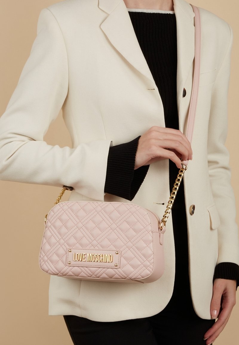 Borsa a tracolla rosa trapuntata con hardware dorato, logo impresso e una tracolla a catena. Presenta una forma rettangolare e una texture in pelle morbida.