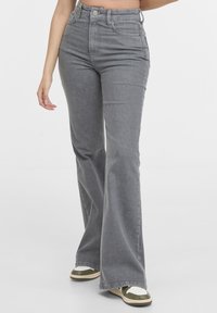 Femme portant un jean gris taille haute à jambes larges et des baskets blanches avec des accents verts, debout devant un fond blanc uni.