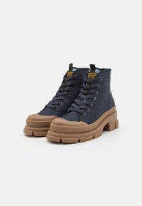 Botas de tobillo de mezclilla azul con suela de goma marrón gruesa, punta redonda y diseño con cordones. Presenta una lengüeta y costuras en contraste.