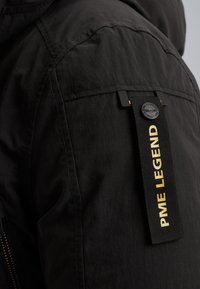 Zwarte jas met een gladde textuur, zichtbare stiksels en een label van stof met "PME LEGEND" in het geel. Ritssluiting aan de zijkant.