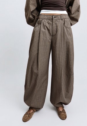 Persona che indossa pantaloni larghi a quadri marroni e neri con vita alta e ballerine marroni, in piedi davanti a uno sfondo chiaro.